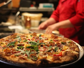 a pizza maker in a red chefs coat makes pizza in a wood fired oven