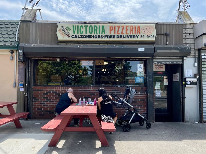 two people and a baby sitting at a red picnic table outside a pizzeria