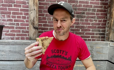 a white mad wearing a red shirt eats a folded slice of pizza while sitting on a wooden bench