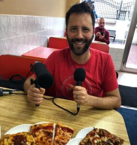 a white man with a beard waring a red shirt holds a microphone while sitting at a table with delicious pizza